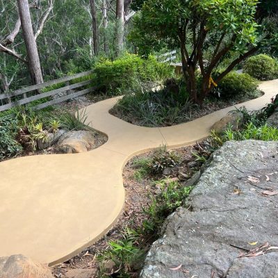 A curving sandstone-colored concrete path surrounded by plants and rocks in a garden setting